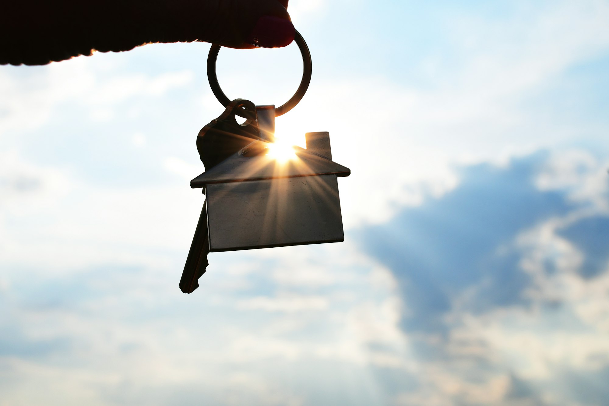 Female hand of realtor holding a set of house keys with home keychain against sky with sunburst